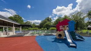 A playground with a slide and climbing structures, surrounded by a fenced area with lounge chairs.