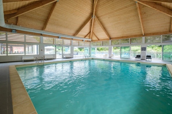 An indoor swimming pool with a wooden ceiling, clear windows, and lounge chairs.