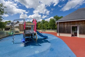 Un parque infantil con tobogán y estructuras de escalada, rodeado de una cerca y un edificio de fachada de madera.