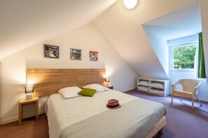Un dormitorio con techo inclinado, una cama con almohadas, una mesita de noche con lámpara, una silla y una ventana con vistas a la vegetación.