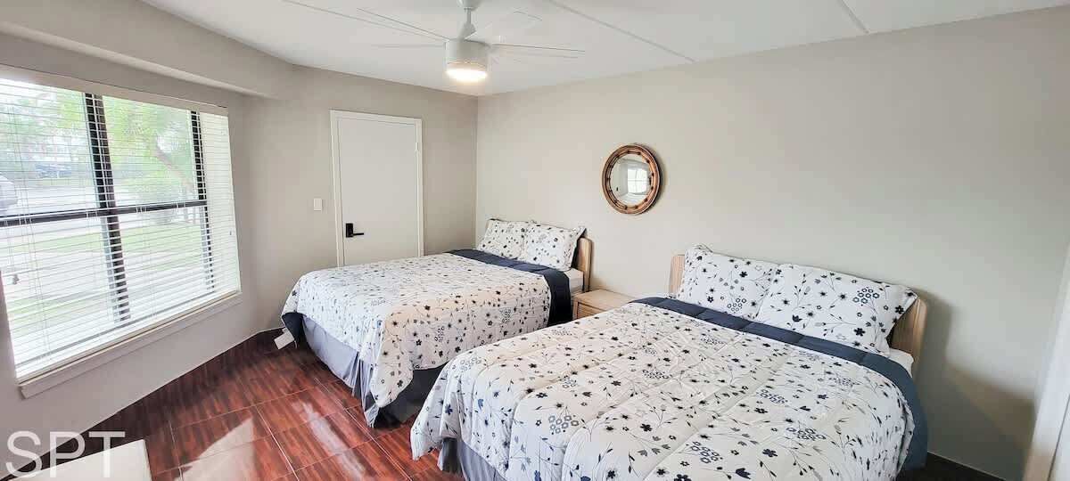 Een slaapkamer met twee bedden, een plafondventilator, een raam met zonwering en een deur.