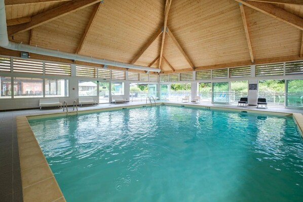 An indoor swimming pool with a wooden ceiling, clear glass windows, and lounge chairs.