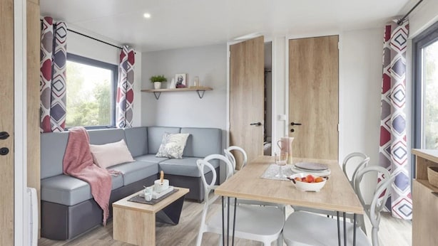 Een modern interieur van een camper, met een grijze bank, witte stoelen, een houten eettafel en gordijnen met een patroon.