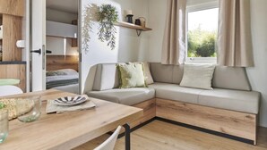 Een moderne, compacte leefruimte met een houten eettafel, een bank met kussens, een hangplant en een raam met gordijnen.