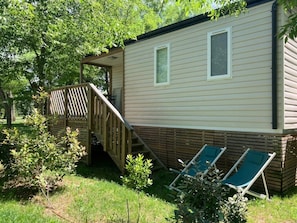 Une maison mobile dotée d’une terrasse en bois, de deux fenêtres et de deux fauteuils de jardin.