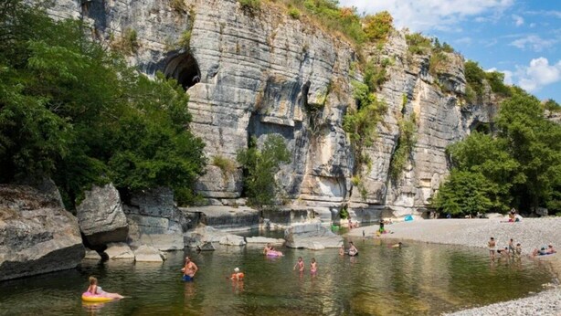 A natural swimming area with rocky cliffs and trees, a clear sky, and people swimming and relaxing.