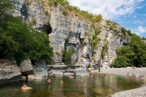 A natural swimming area with rocky cliffs and trees, a clear sky, and people swimming and relaxing.