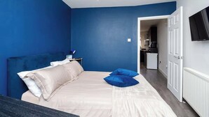 Un dormitorio con cama, almohadas, una mesita de noche, televisor y vista a la cocina.