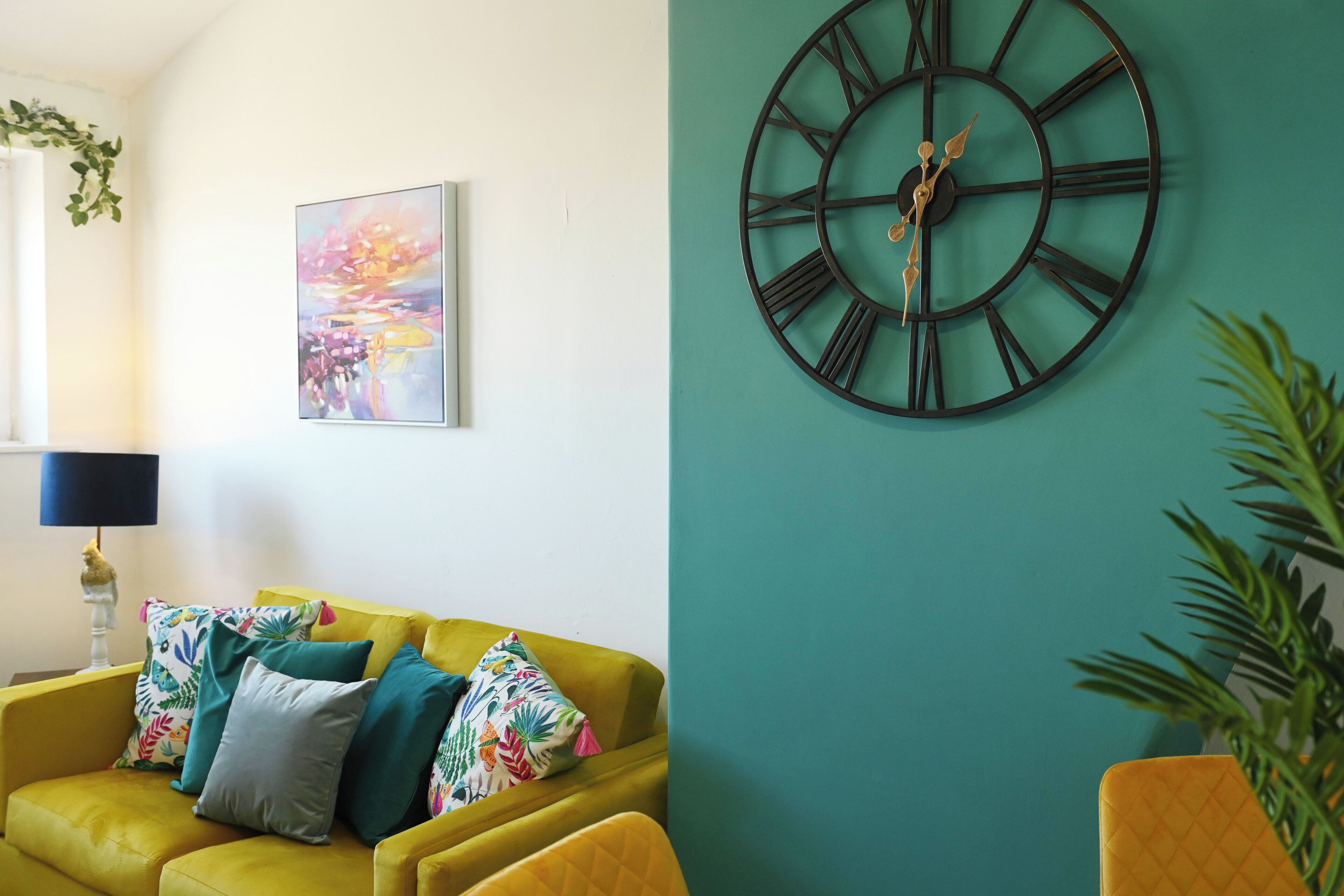 Une pièce avec un canapé jaune, une grande horloge murale et un tableau abstrait aux couleurs vives.