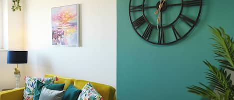 A room with a yellow sofa, a large wall clock, and a colorful abstract painting.
