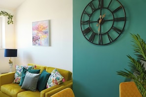 A room with a yellow sofa, a large wall clock, and a colorful abstract painting.