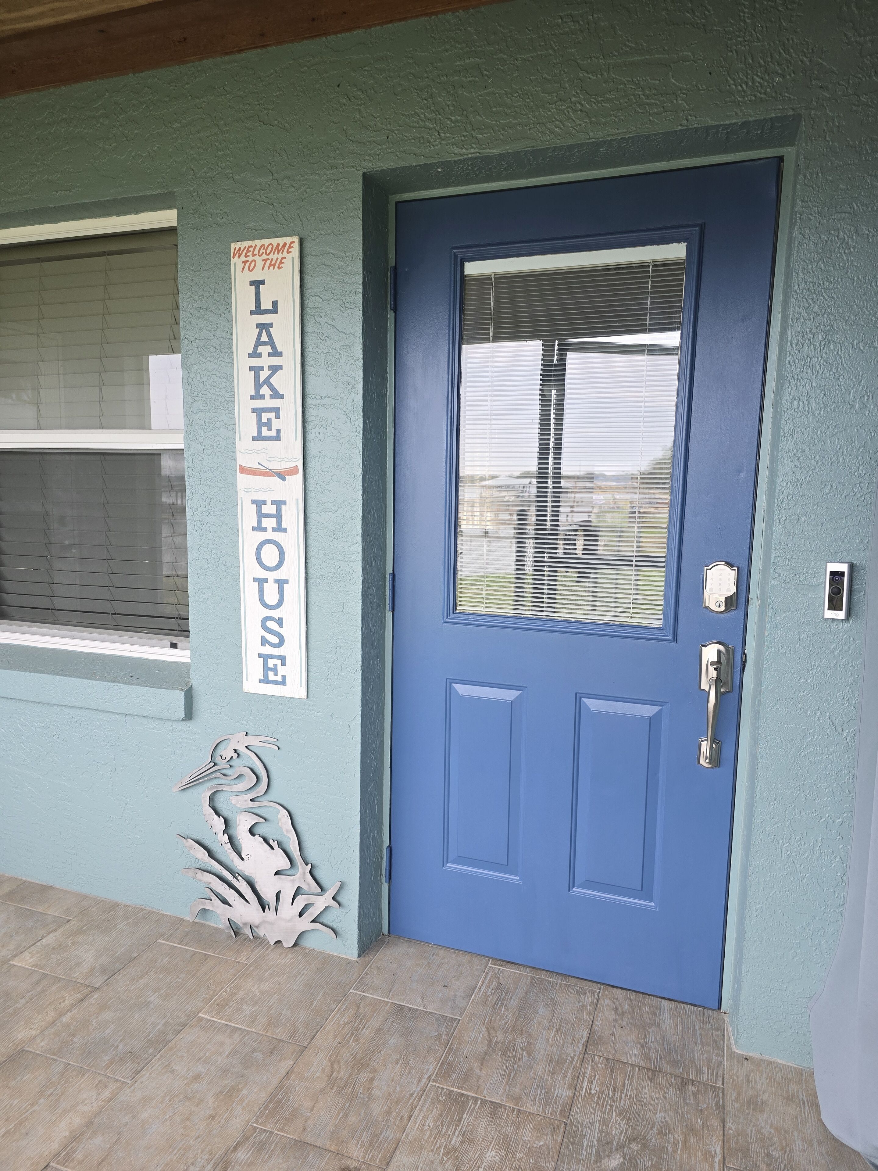 Eine blaue Tür mit Fenster, ein Schild mit der Aufschrift 'Welcome to the Lake House' und eine Vogelplastik an der Wand.