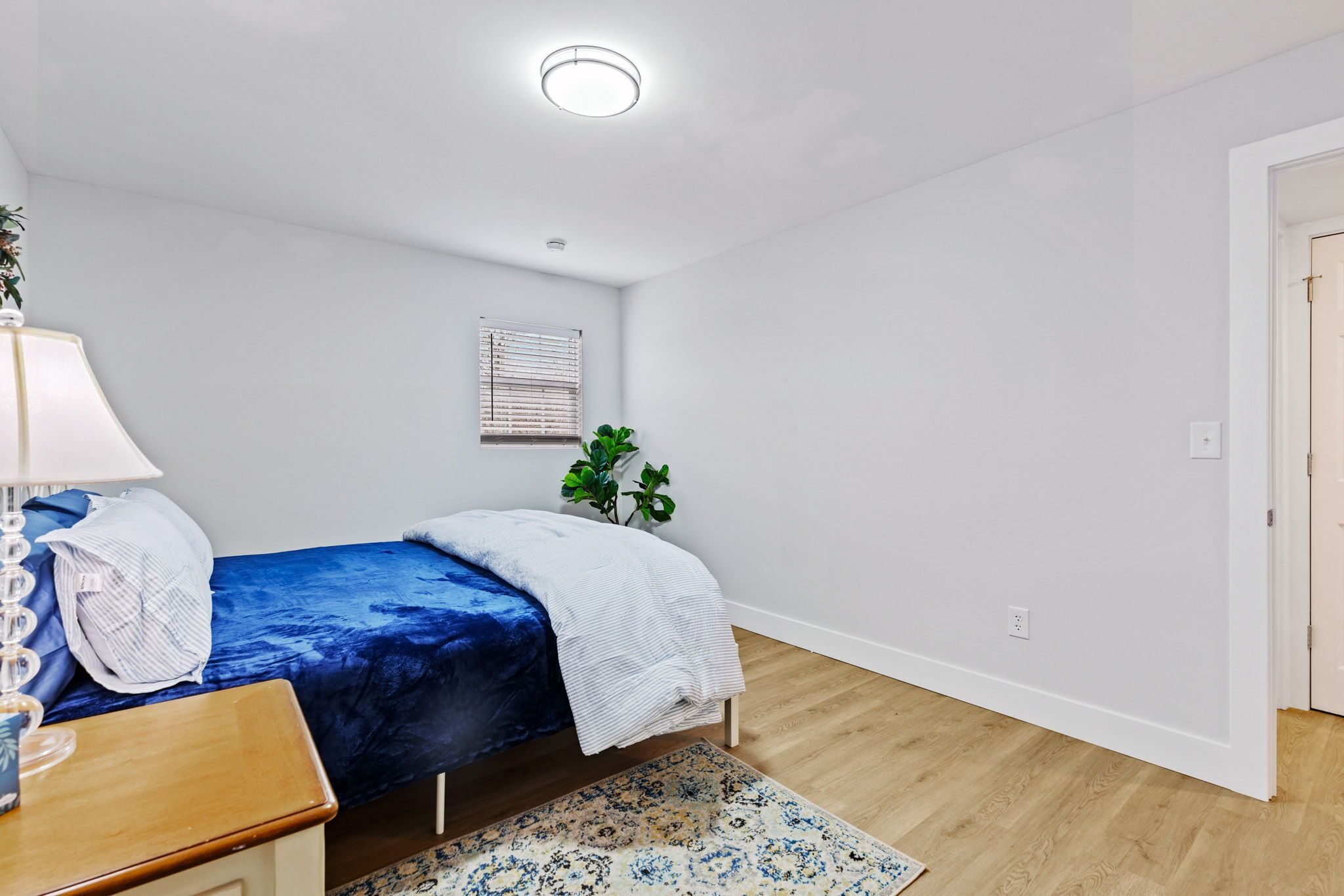 A bedroom with a bed, a bedside table, a lamp, a window with blinds, a plant, and a wooden floor.