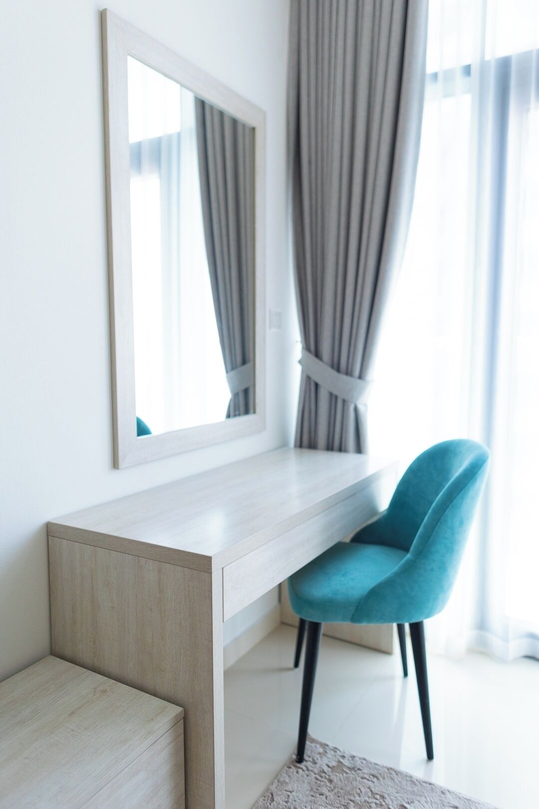 Quarto moderno com escrivaninha de madeira clara, cadeira azul, espelho grande e cortinas cinzentas.