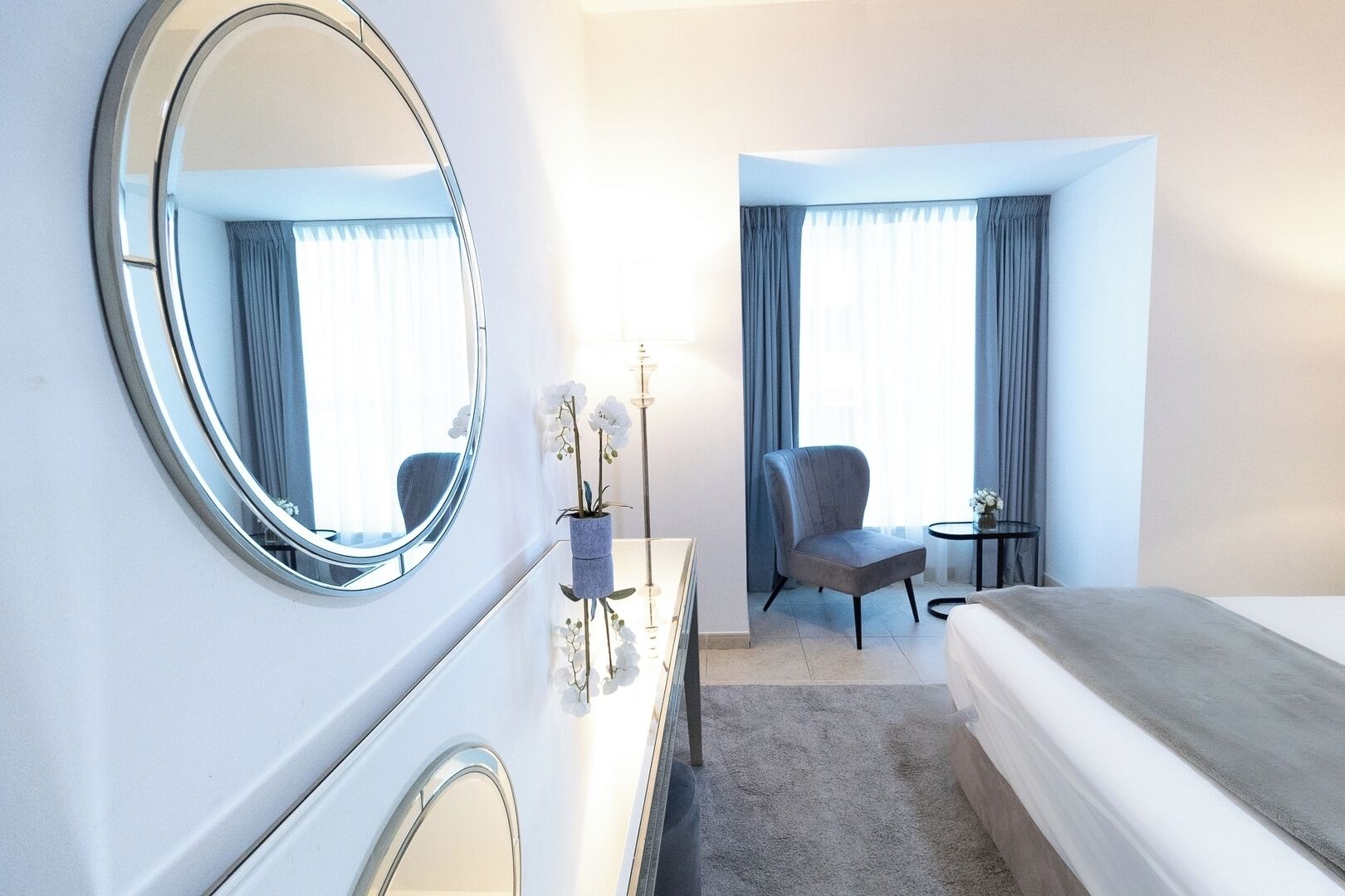 Une chambre d’hôtel avec un grand miroir rond, un lit, une chaise, une petite table et une commode.