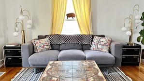 Ein Wohnzimmer mit grauem Sofa, gemusterten Kissen, einem Couchtisch mit Rundmuster und einem Fenster mit gelben Vorhängen.