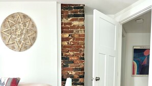 Ein Schlafzimmer mit einer Ziegelwand, einem Bett mit einem gemusterten Kissen, einem Nachttisch mit einer Lampe, einem Teppich und einer Tür zu einem anderen Raum.