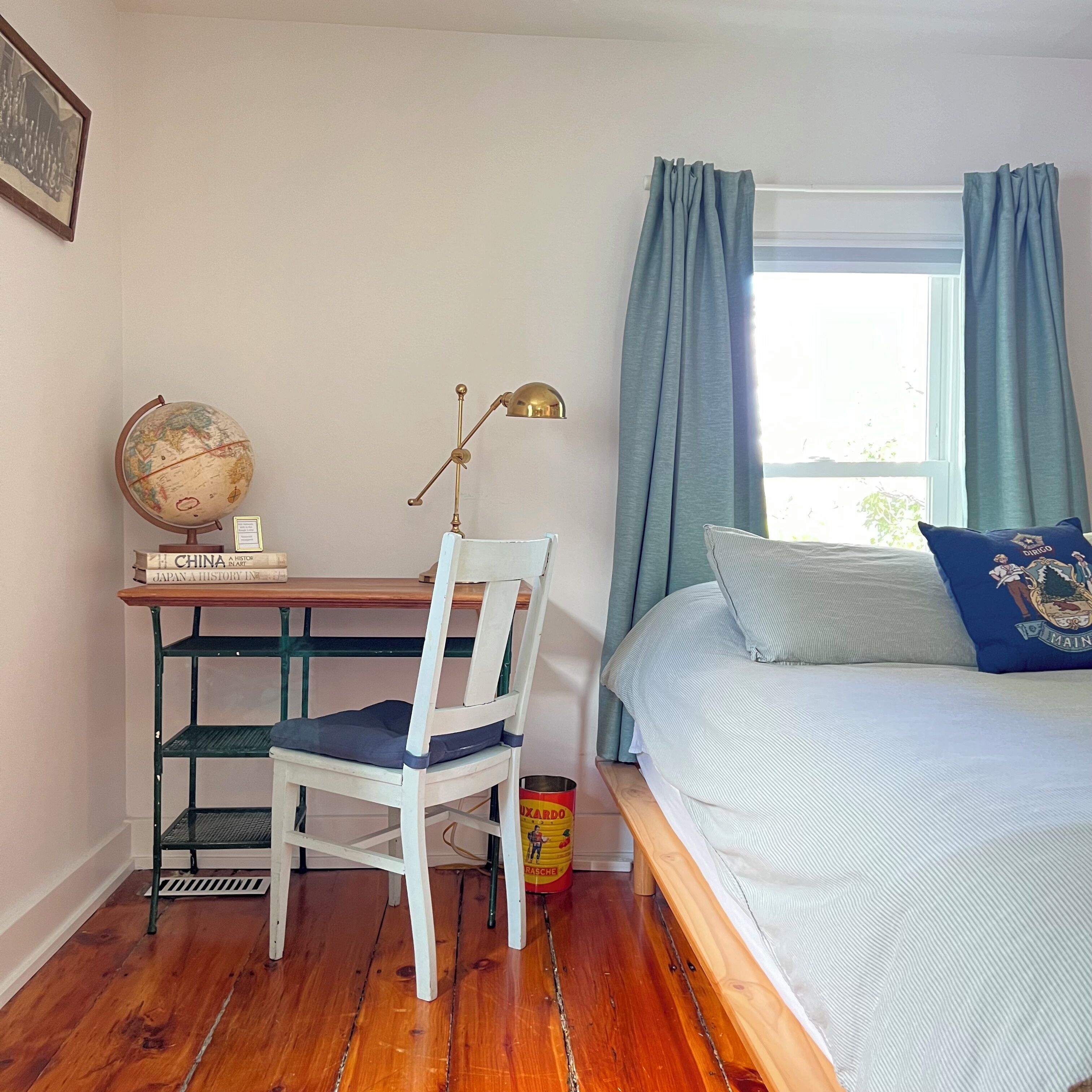 A bedroom with a bed, a desk with a globe and books, a chair, and a window with curtains.
