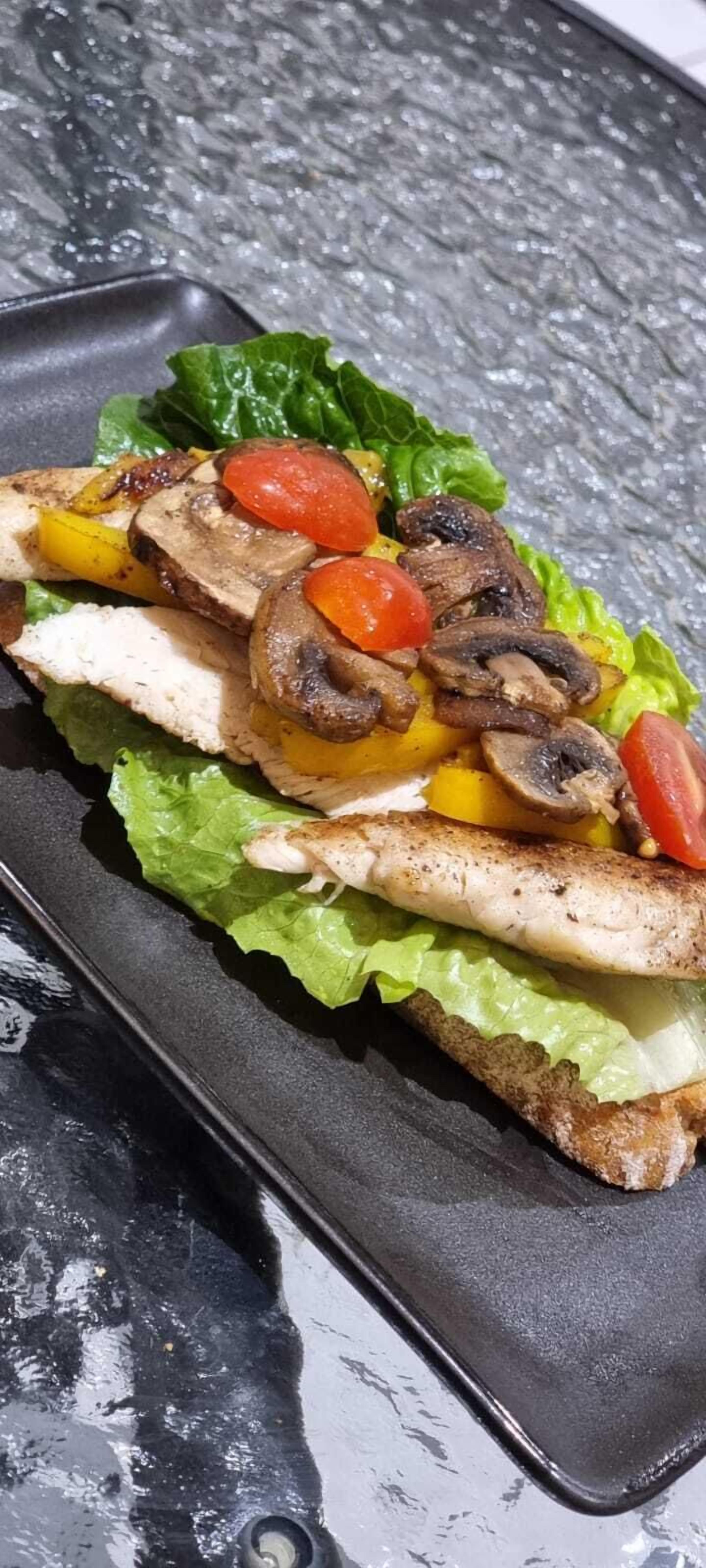 A sandwich on a black plate with lettuce, grilled chicken, mushrooms, and tomatoes.