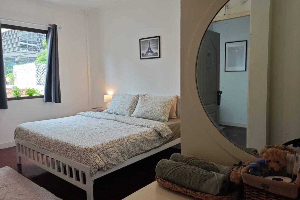 A bedroom with a bed, a bedside table, a lamp, a window with curtains, a framed picture on the wall, a round mirror, and a basket with toiletries.