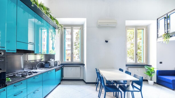 Une cuisine moderne avec des meubles de couleur turquoise, une table à manger blanche et des chaises bleues. Deux fenêtres laissent entrer la lumière naturelle, et un canapé bleu se trouve dans l’espace salon.