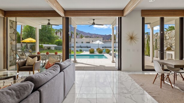 A modern living room with a large gray sofa, a dining table with chairs, and a view of a swimming pool and mountains in the background.