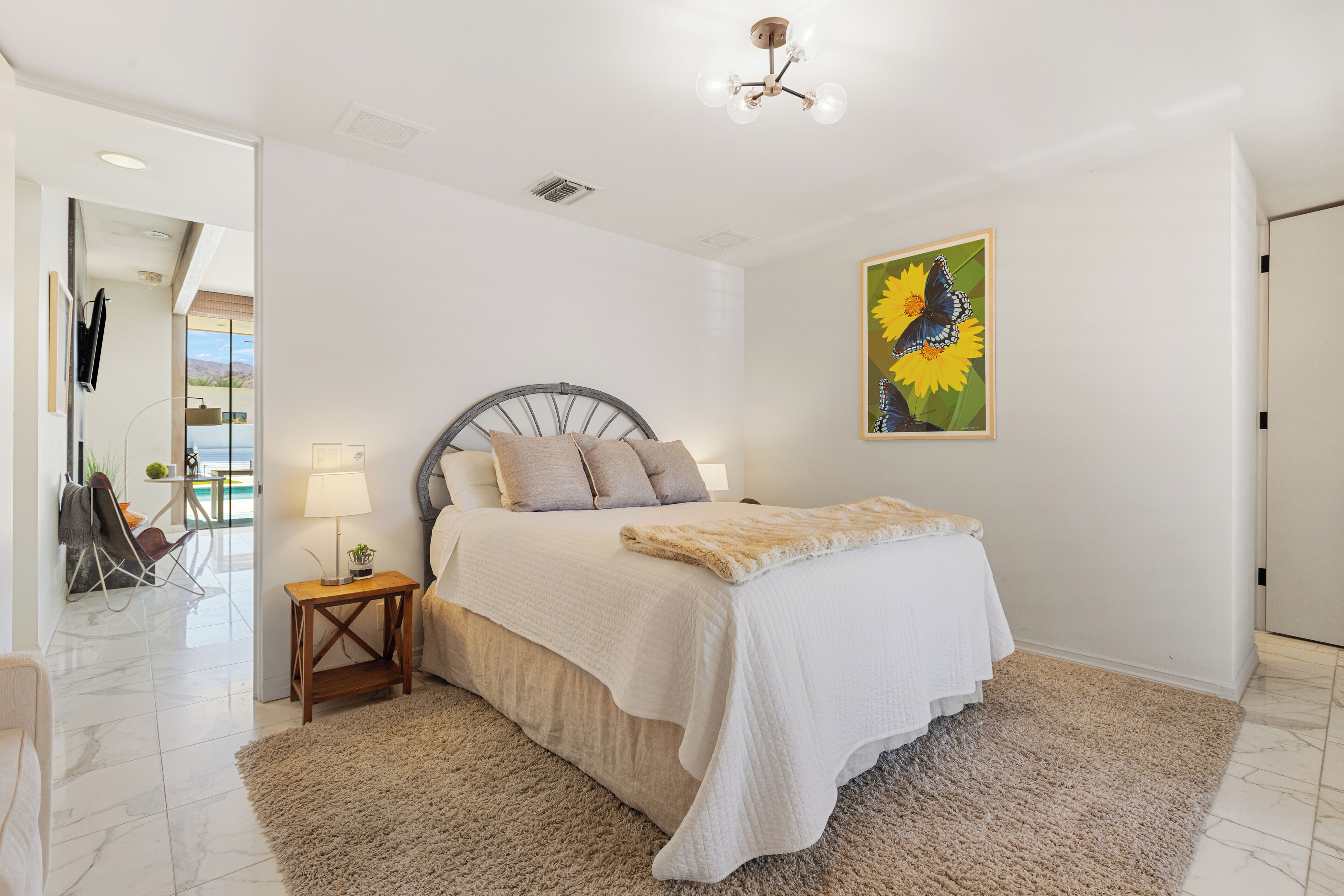 A bedroom with a bed, a nightstand, a lamp, a painting, and a view of the outside.