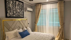 Un dormitorio con cama, cabecera, aire acondicionado de pared y ventana con cortinas traslúcidas.