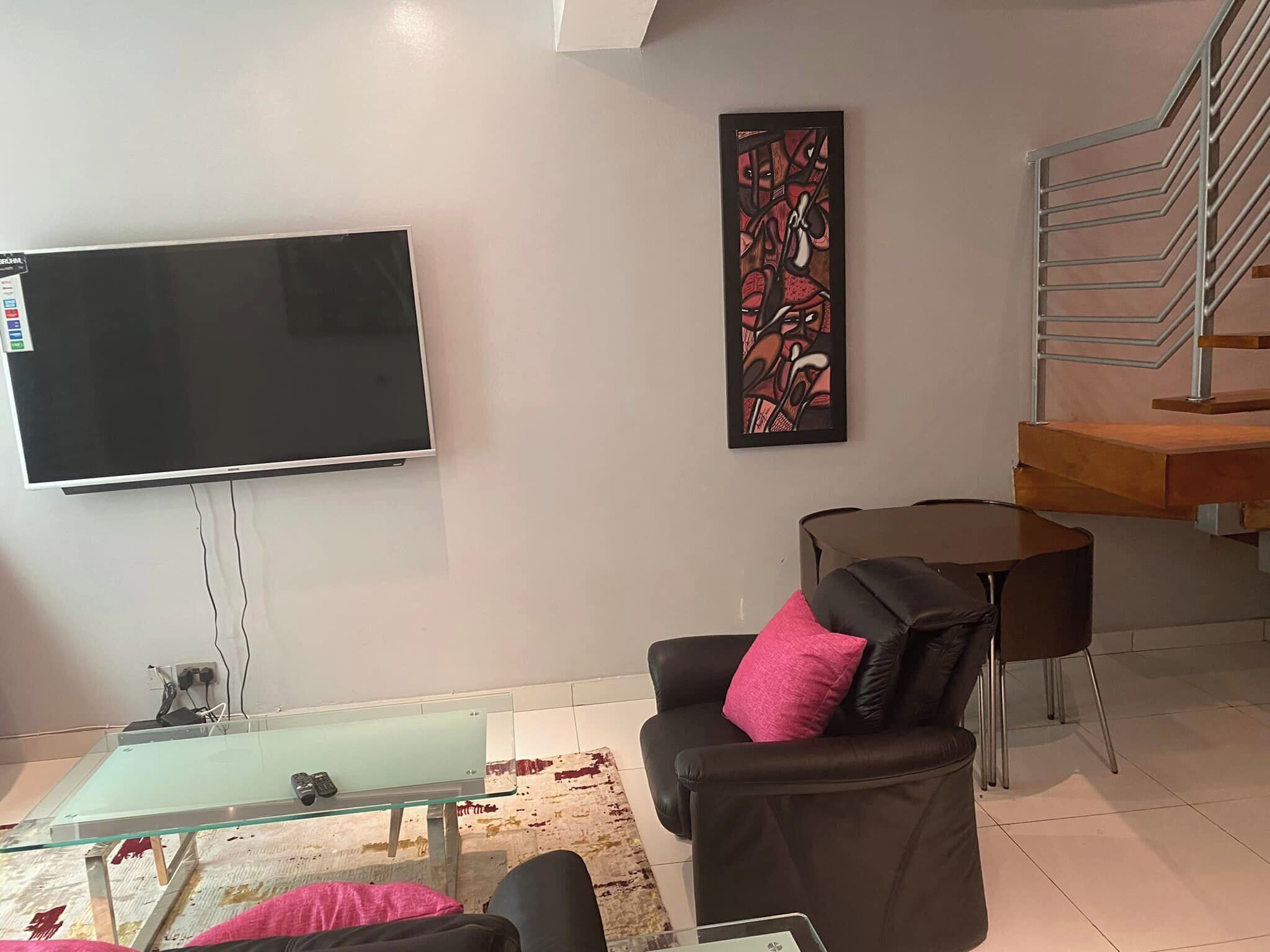 En moderne stue med et fladskærms-tv, et glasbord, en sort lædersofa med en pink pude, et lille rundt bord og en trappe med trægelænder.