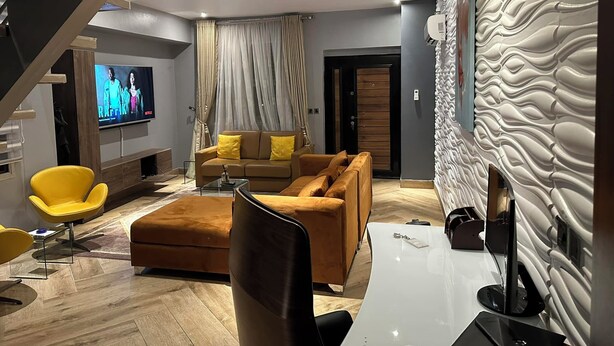 Una sala moderna con un sofá, reposapiés y televisor. Se ve una escalera a la izquierda y una pared texturizada a la derecha.