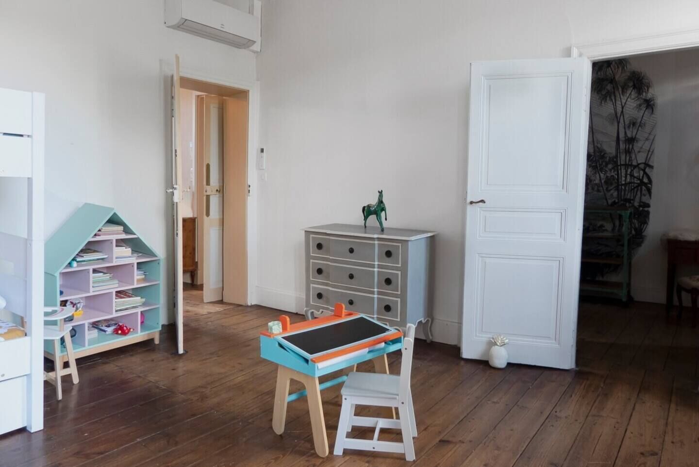 Ein Kinderzimmer mit Holzboden, einem Schreibtisch mit Stuhl, einem Bücherregal und einer Kommode mit Schubladen.