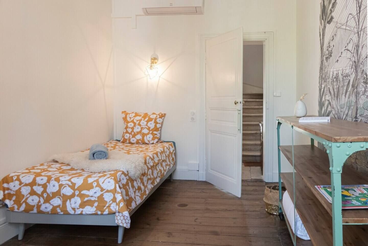 Ein Schlafzimmer mit Bett, Nachttisch, Wandleuchte, einer Tür zu einer Treppe und einer Wand mit einem Pflanzenmotiv.
