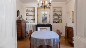 Comedor con una mesa redonda, sillas, una lámpara de araña, chimenea y estanterías con diversos objetos.