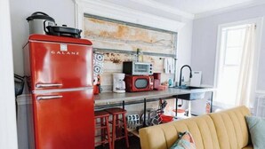 A compact kitchen with a red Galanz refrigerator, a microwave, and a coffee maker. There is a yellow sofa with a colorful pillow in the living area. - Cozy Midtown Mobile Home; Fireplace & Pet-Friendly (AL)