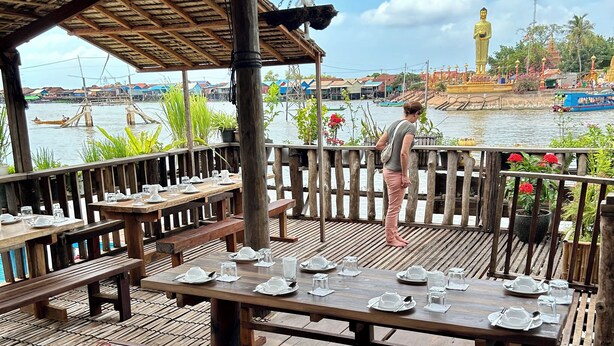 Een houten terras buiten met tafels en banken, met uitzicht op een waterpartij met boten en een groot gouden beeld op de achtergrond.