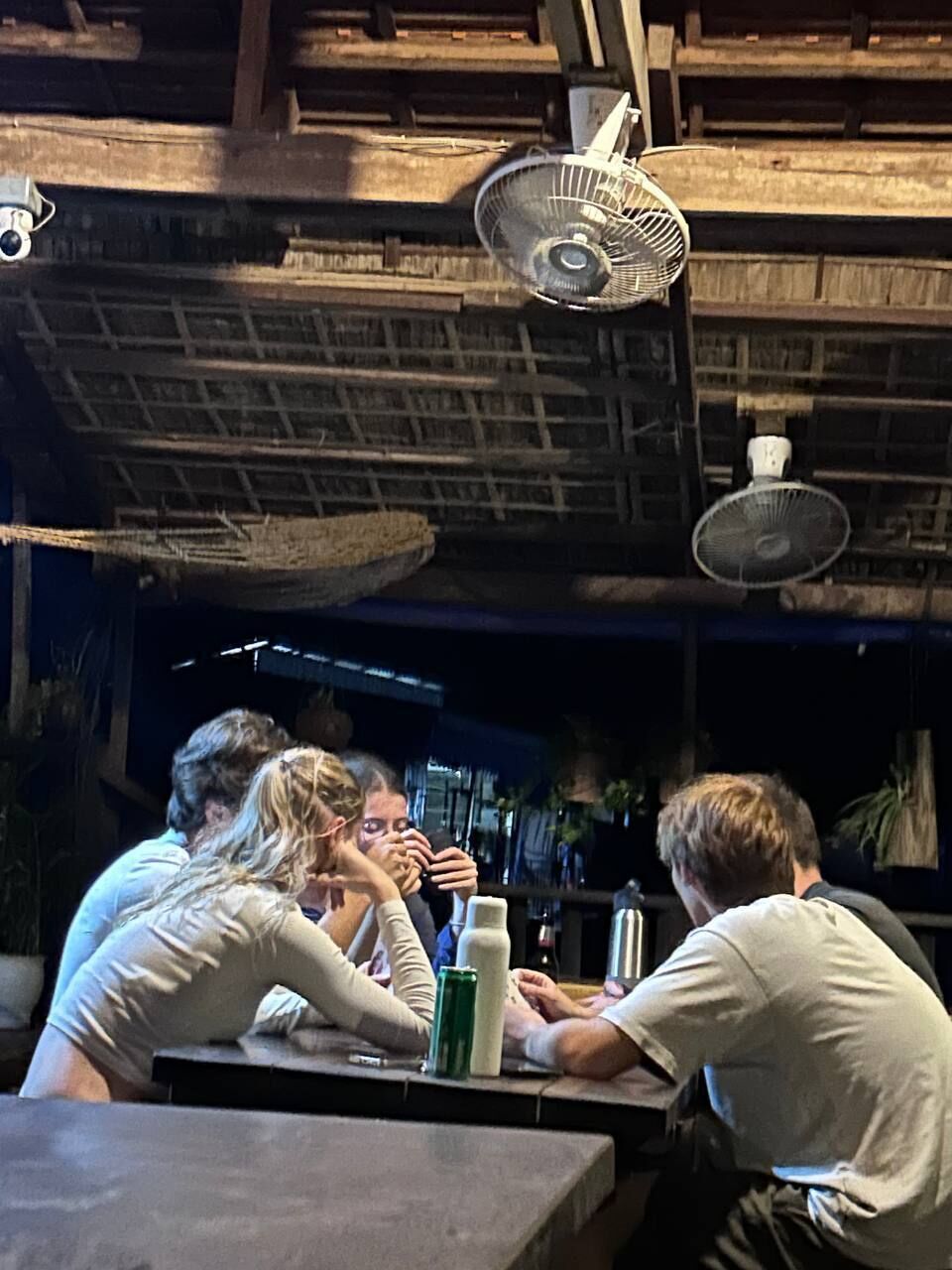 A group of people sitting at a table in a rustic setting with fans overhead.