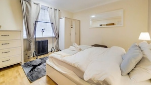 A bedroom with a bed, a dresser, a radiator, a window with curtains, and a lamp. - Quebec Residence (London)