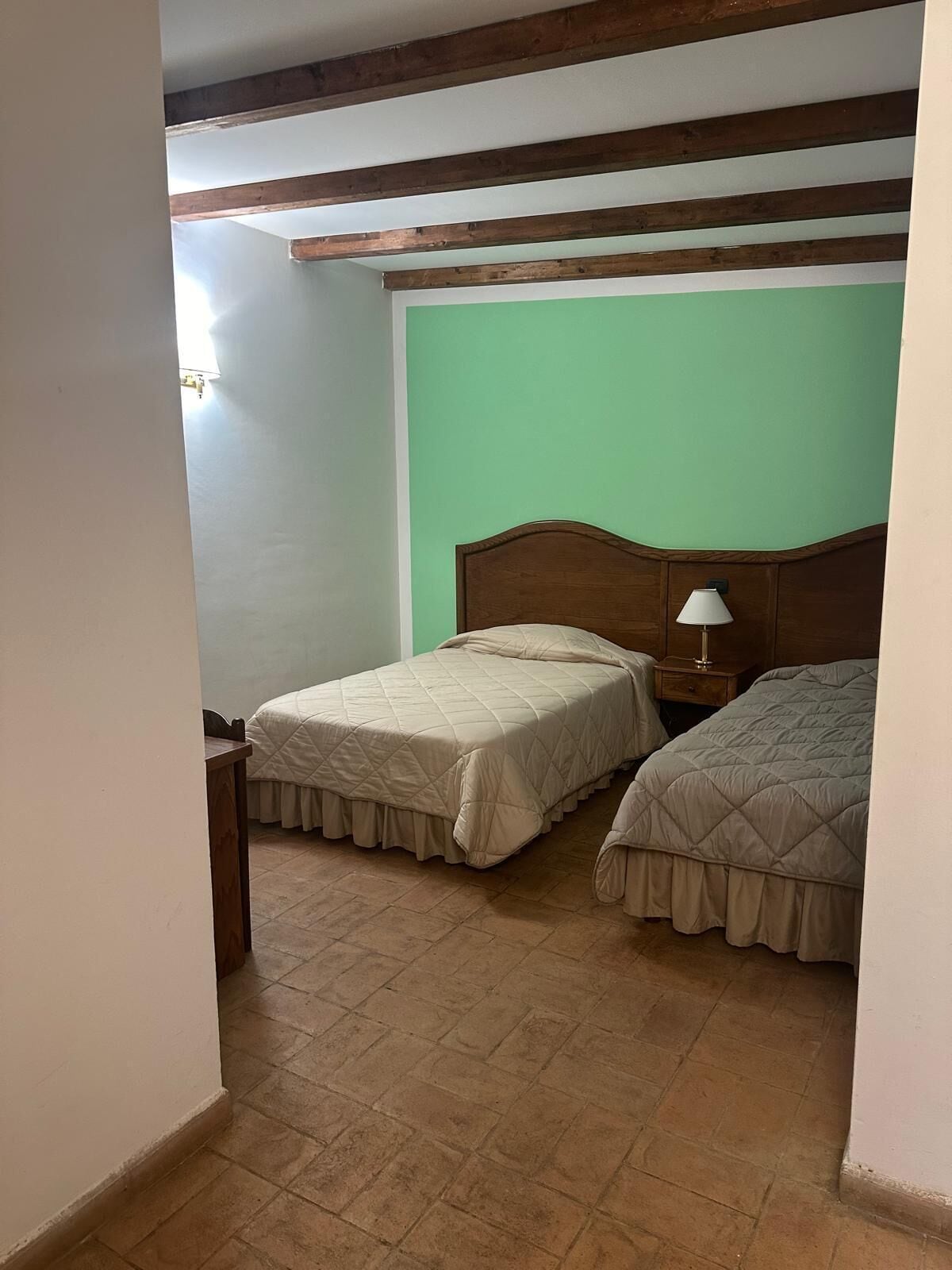 Une chambre avec deux lits, des poutres en bois au plafond, un mur vert et une table de chevet avec une lampe.