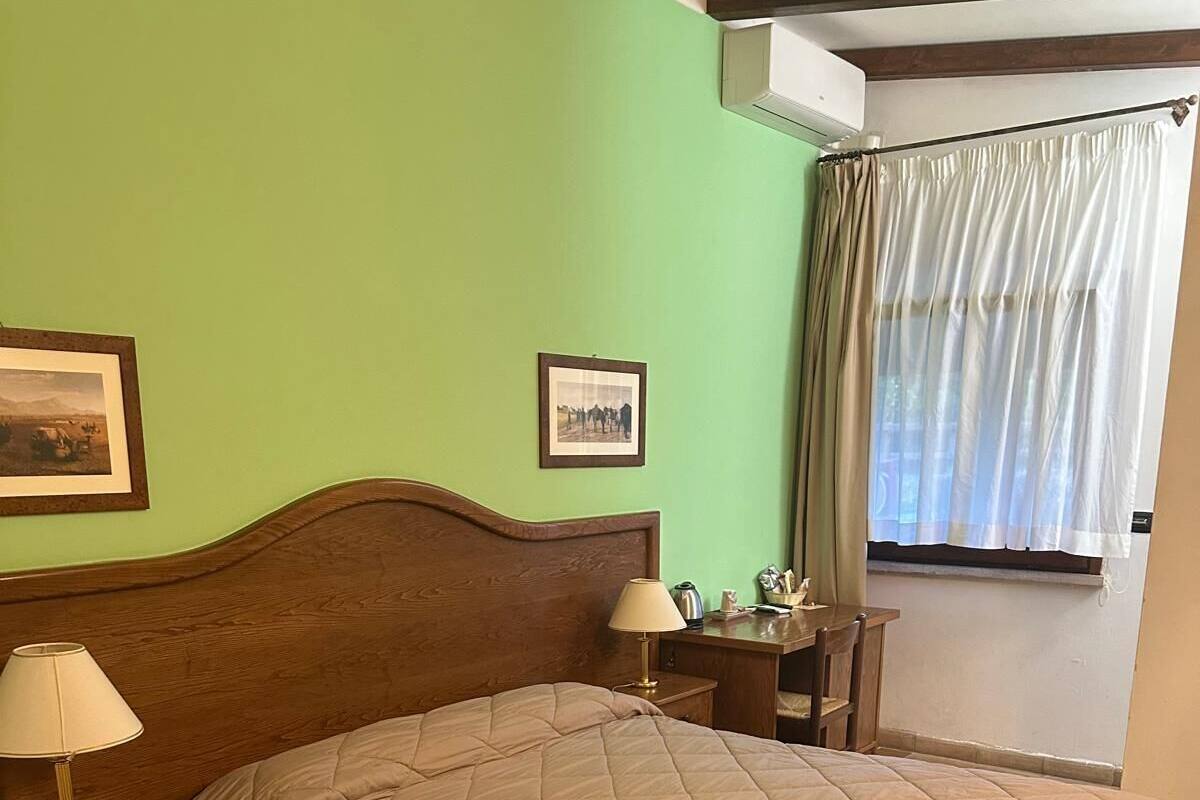 Een slaapkamer met een houten hoofdbord, een bed met een beige sprei, twee nachtkastjes met lampen, een klein bureau en een raam met gordijnen.
