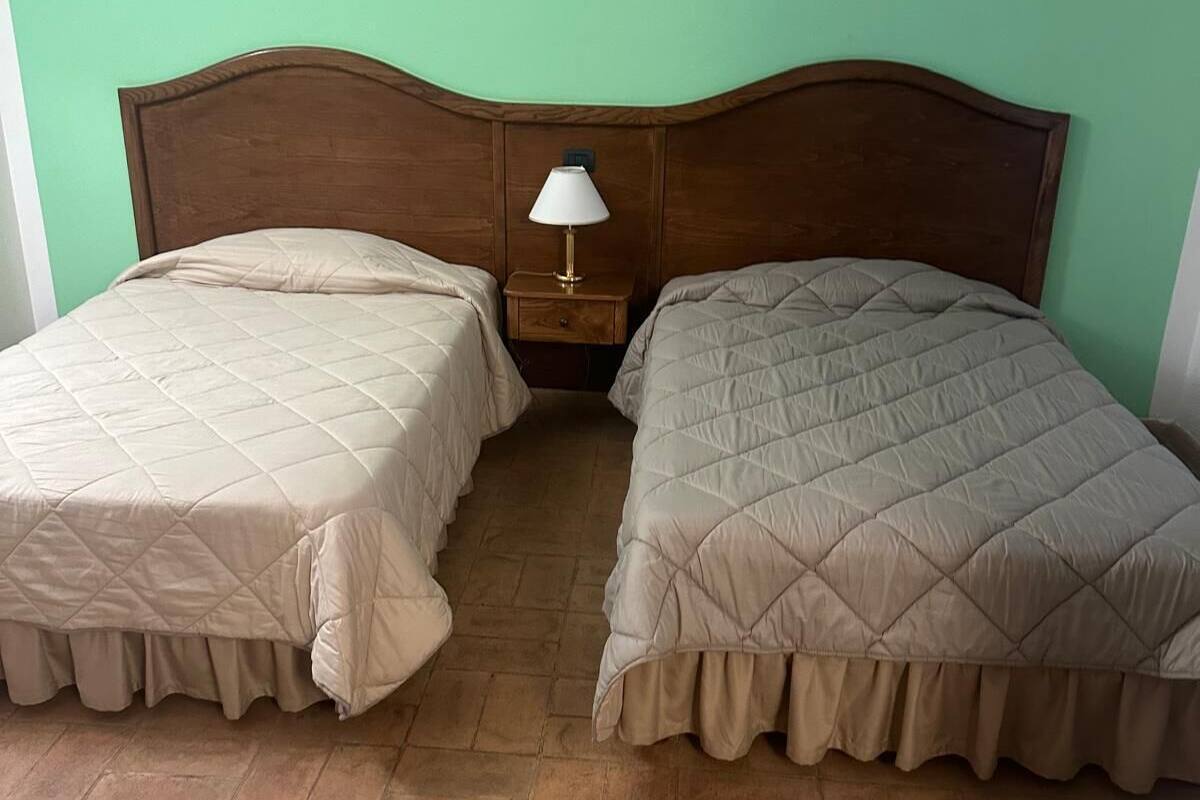 Een kamer met twee bedden, elk met een houten hoofdbord en een beige gewatteerde spreideken. Daartussen bevindt zich een klein houten nachtkastje met een witte lamp.