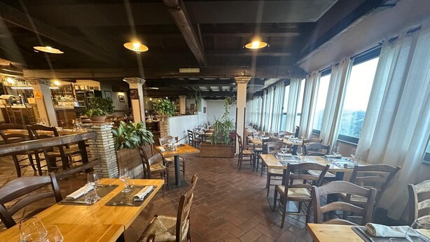 A restaurant with wooden tables and chairs, potted plants, and large windows.