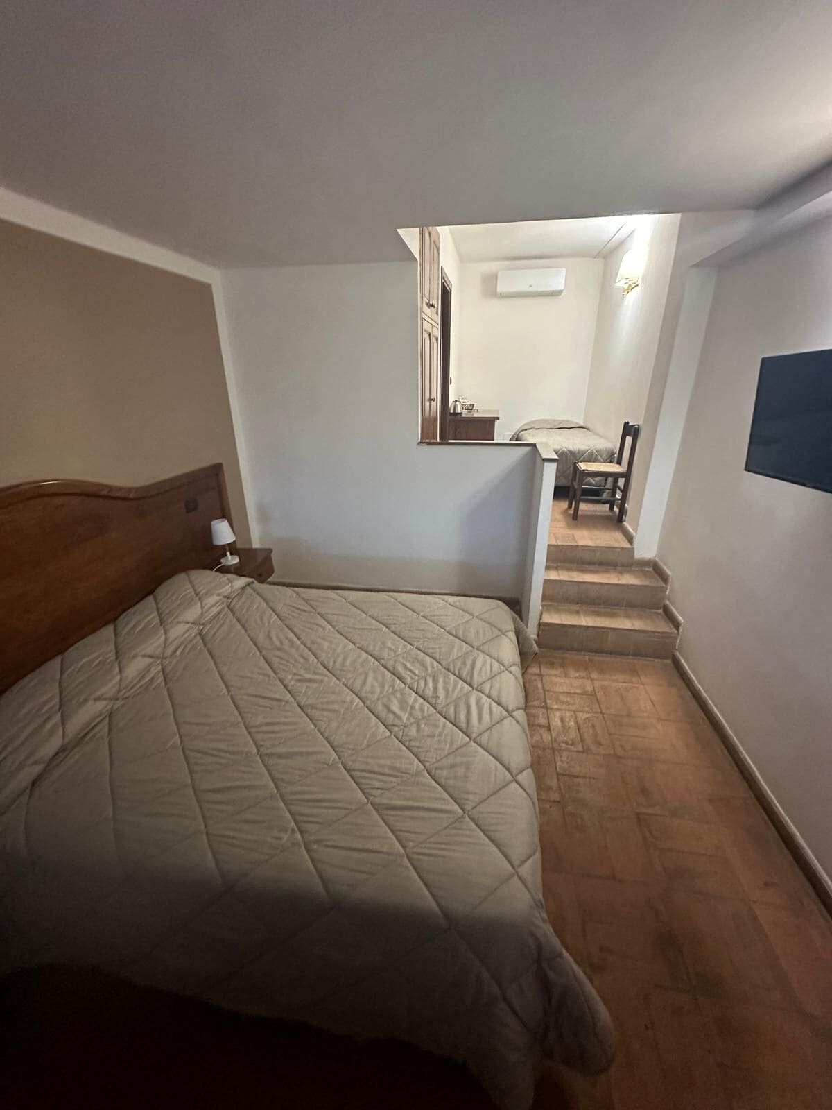 Een slaapkamer met een bed, een nachtkastje, een stoel en een kleine tafel met een lamp. Er is een trap die leidt naar een badkamer en een keukenhoek.