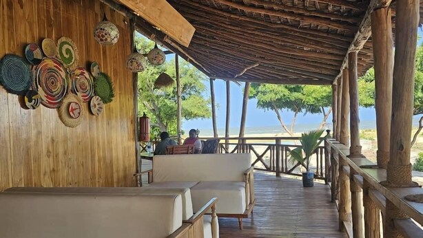 Una terraza de madera con asientos acolchados blancos, un techo de paja y vista a la playa.