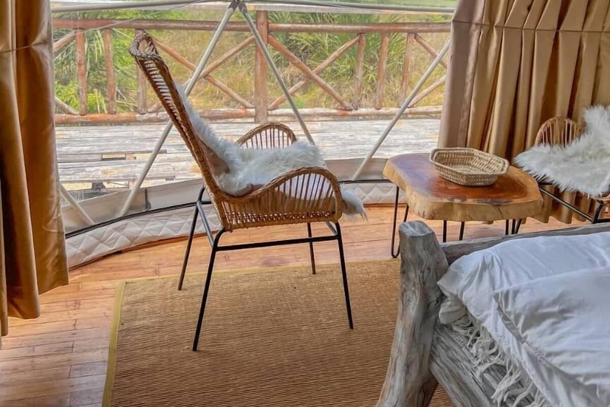 Une pièce aménagée sous une tente, avec un lit, une chaise en osier, une petite table sur laquelle se trouve un panier, et une vue sur le paysage.