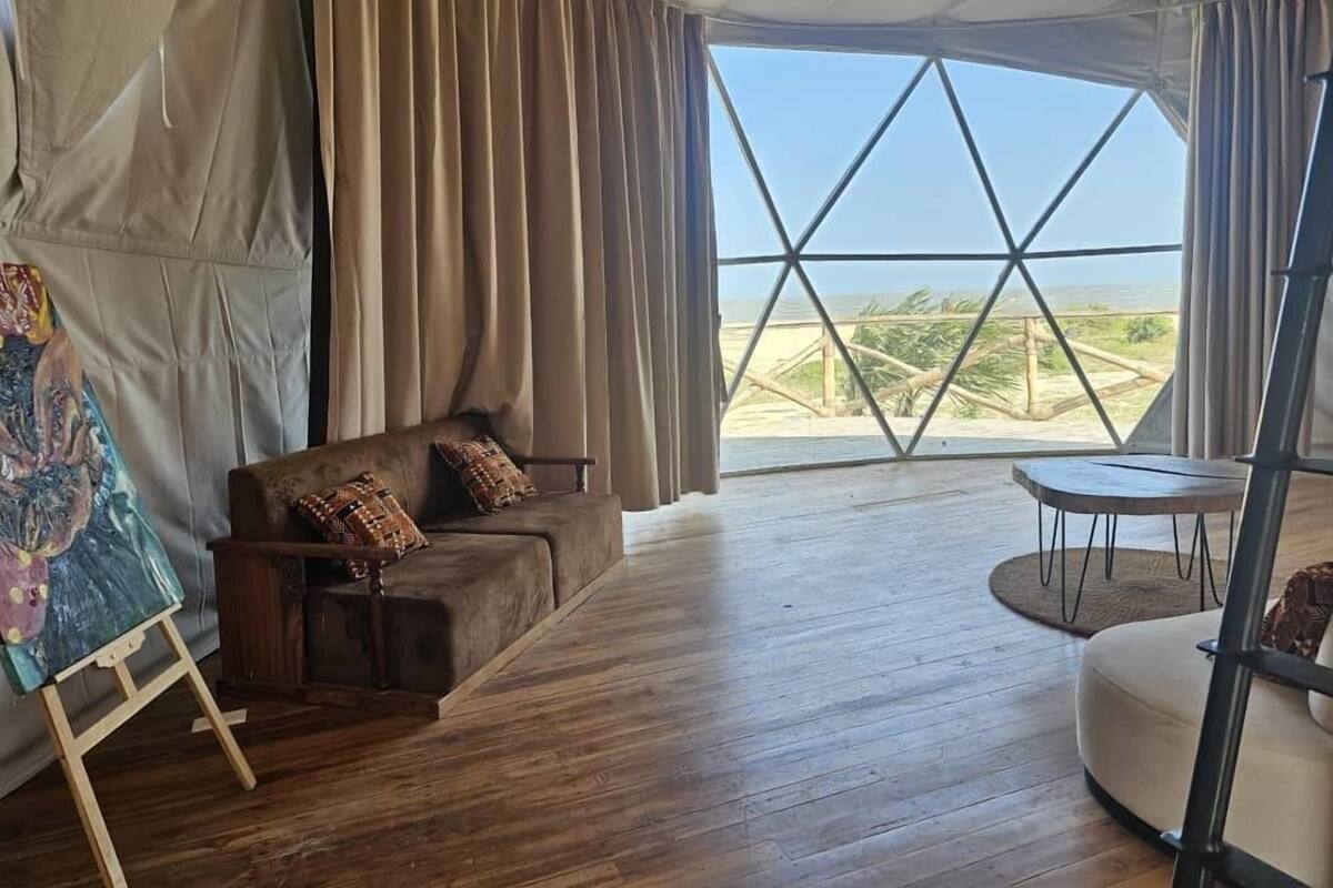 Une pièce aménagée comme une tente, avec un plancher en bois, un canapé agrémenté de coussins à motifs, une petite table et une grande fenêtre donnant sur la plage.