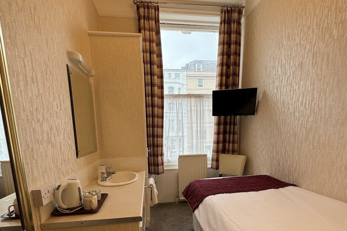 A hotel room with a bed, a bedside table, a mirror, a television, a window with curtains, and a bathroom with a sink and a kettle.
