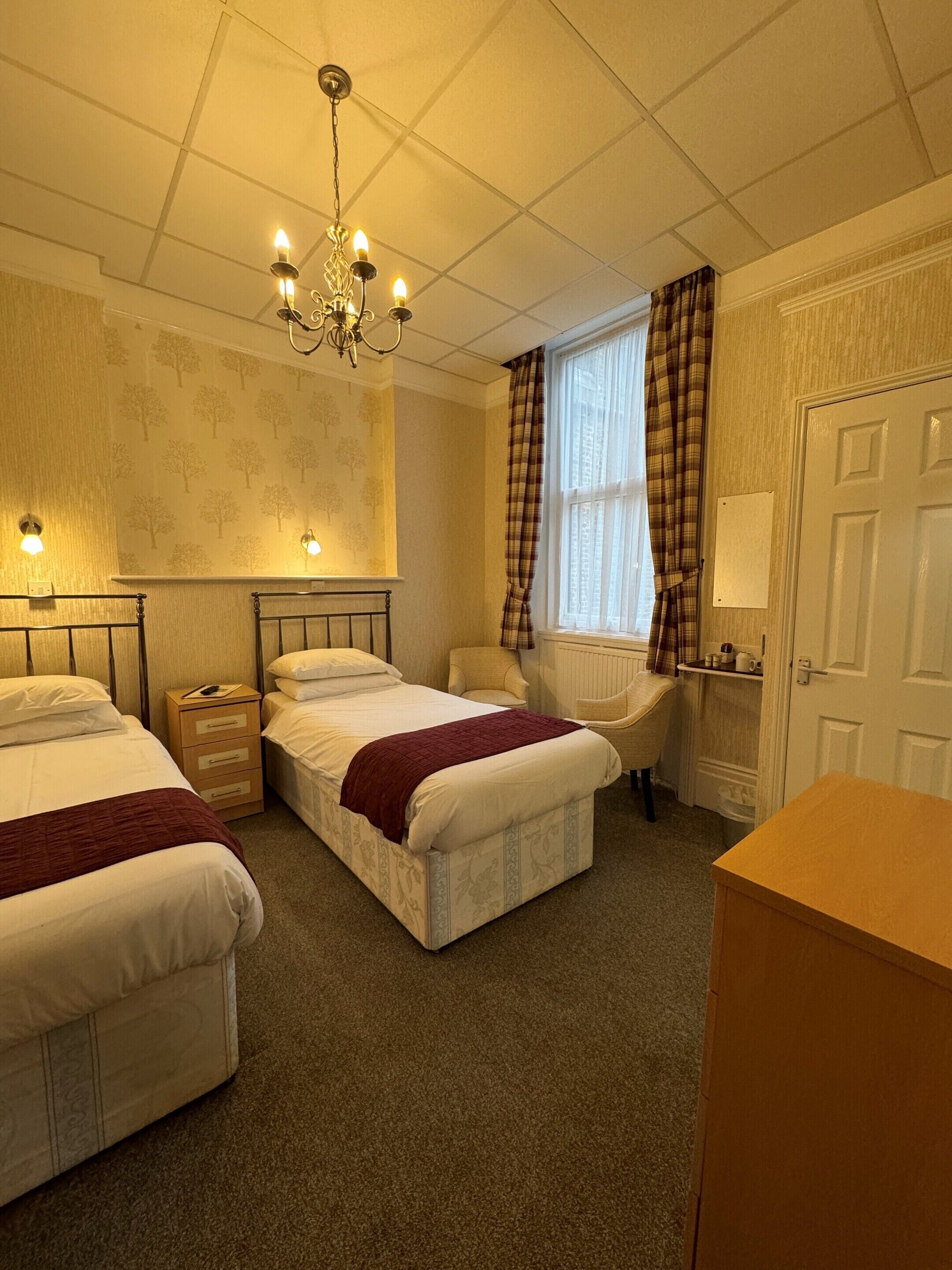 A hotel room with two beds, a chandelier, a window with curtains, a chair, and a desk.