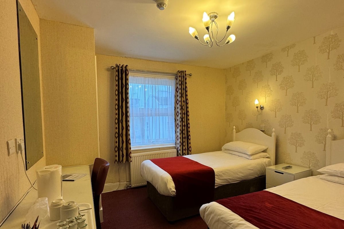 A hotel room with two beds, a desk with toiletries, a mirror, and a window with curtains.