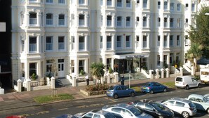 Un hôtel à plusieurs étages, de style architectural classique, avec une entrée principale dotée d’un escalier, un espace de stationnement et une vue sur la rue où sont garées des voitures.