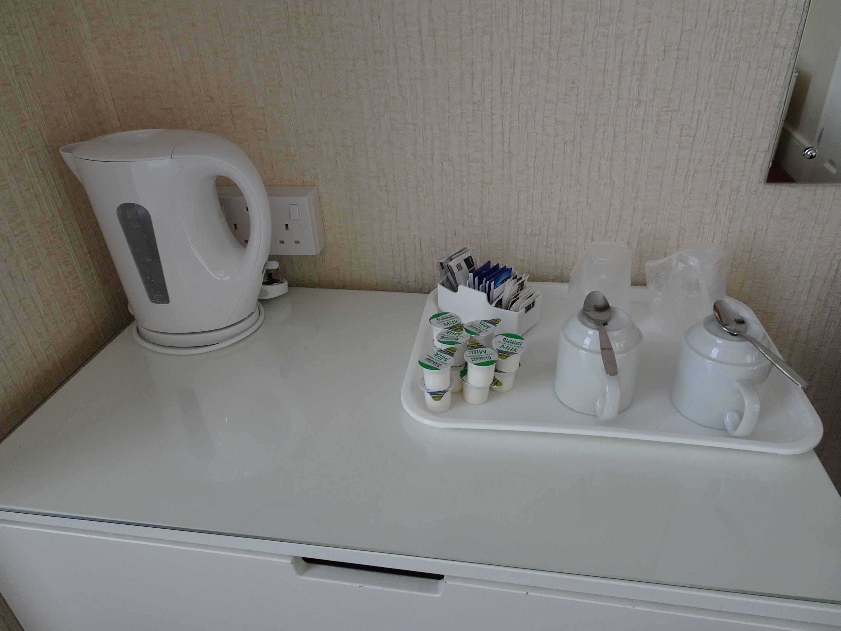 Un mini bar blanco con una tetera, leche en envases y una bandeja con cubiertos.